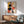 May include: Abstract geometric art print in wooden frame hanging above mid-century modern credenza. Artwork features large black circles, orange and red shapes, blue rectangles, and yellow elements in Bauhaus style. Room has white walls with dramatic sunlight shadows, teal upholstered chair with wooden arms, white ceramic table lamp, and partial view of round coffee table.