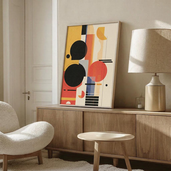 May include: Abstract geometric poster with colorful circles, rectangles and shapes in yellow, orange, red, black and blue displayed in thin frame on wooden sideboard. Modern minimalist room setting with cream textured chair, ceramic table lamp with beige shade, small wooden stool, and neutral beige walls.
