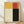 May include: Framed abstract art print with four geometric color blocks - yellow, red-orange, cream, and black arranged in a grid pattern. Light wood frame sits on wooden floor against white wall with natural shadows. Minimalist modernist style artwork display in bright interior setting.