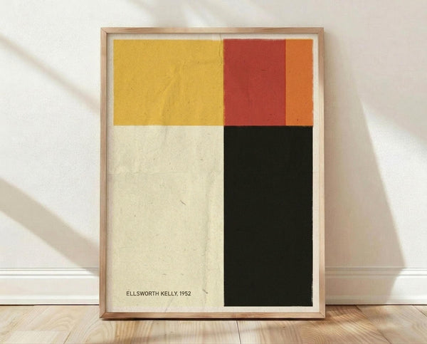 May include: Framed abstract art print with four geometric color blocks - yellow, red-orange, cream, and black arranged in a grid pattern. Light wood frame sits on wooden floor against white wall with natural shadows. Minimalist modernist style artwork display in bright interior setting.
