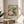 May include: Framed poster featuring a green hummingbird in flight surrounded by pink and white peonies with lush foliage, displayed on a round wooden side table next to a beige upholstered chair. Natural sunlight streams across the neutral-toned room setting, casting soft shadows on the white wall behind the vintage-style botanical artwork in a thin wooden frame.