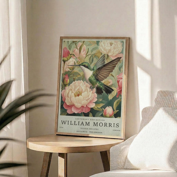 May include: Framed poster featuring a green hummingbird in flight surrounded by pink and white peonies with lush foliage, displayed on a round wooden side table next to a beige upholstered chair. Natural sunlight streams across the neutral-toned room setting, casting soft shadows on the white wall behind the vintage-style botanical artwork in a thin wooden frame.