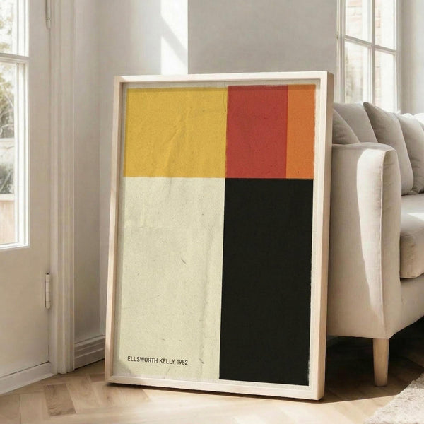 May include: Abstract geometric art print in light wood frame leaning against white wall next to beige upholstered sofa. Artwork features four color blocks - golden yellow, cream white, red-orange, and dark green sections with "ELLSWORTH KELLY, 1952" text in corner. Bright natural light streams through tall windows onto hardwood floors in minimalist living room setting.