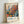 Framed vintage Japanese exhibition poster featuring abstract botanical artwork with colorful circular forms in orange, blue, yellow, and white resembling stylized flowers and seed pods against a soft gradient background. The poster displays Japanese characters and "JAPAN 1984" with "MUSÉE D'ART MODERNE DE LA VILLE DE PARIS" at bottom, dated "5. JUIN - 19. JUIN 1986" at top. Light wood frame leaning against white wall corner.
