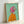 May include: Framed painting of an Italian Greyhound wearing a bright pink headscarf and orange coat, painted in a portrait style against a mint green background. The artwork is displayed in a natural wood frame leaning against a white wall on a light surface, showing visible paint texture and brushstrokes.