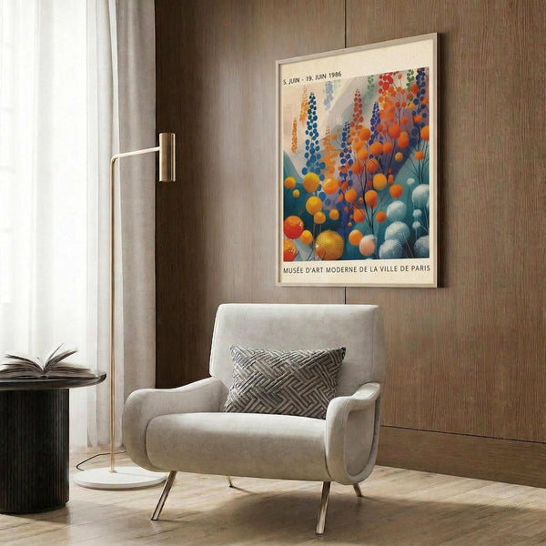 May include: Colorful botanical poster with blue, orange, and teal spherical flowers displayed on wood-paneled wall above beige upholstered armchair with geometric throw pillow. Scene includes gold floor lamp and dark round side table with open book. Poster shows exhibition details for Musée d'Art Moderne de la Ville de Paris dated June 1986.