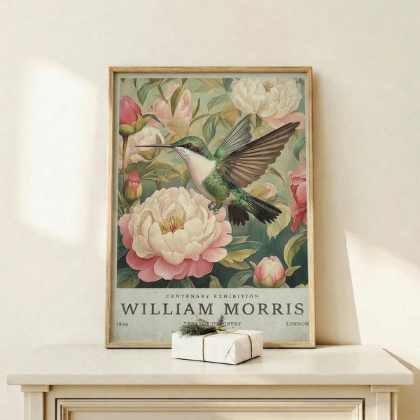 May include: Framed botanical poster featuring a green and white hummingbird with spread wings hovering near large pink and white peonies with green foliage. Text reads "Centenary Exhibition William Morris 1934 London" at bottom. Natural wood frame sits on light wooden surface against cream wall, with small wrapped white gift box nearby.