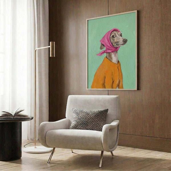 May include: Framed poster print showing an Italian Greyhound wearing a pink headscarf and orange sweater against a mint green background, displayed on wood paneled wall above a beige upholstered armchair with geometric patterned pillow, alongside a brass floor lamp and side table with open book in a modern living room setting.