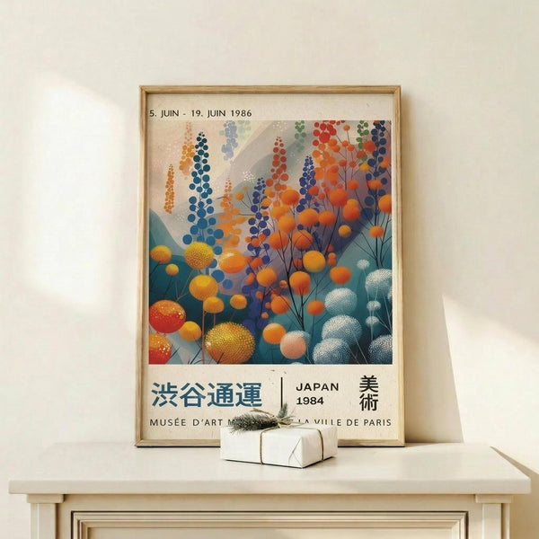 May include: Framed vintage Japanese exhibition poster displaying colorful abstract botanical artwork with circular flower forms in orange, blue, and teal tones, featuring Japanese text and "JAPAN 1984" with museum attribution. Light wood frame sits on minimalist white wooden shelf beside small wrapped gift box with evergreen sprig against cream wall.