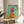 May include: Framed artwork displayed on a round wooden side table next to a beige upholstered chair with white pillow. The painting shows an Italian Greyhound dog wearing a bright pink headscarf and orange sweater against a mint green background. Natural lighting creates soft shadows on white walls, with a plant visible in the corner of the modern, minimalist room setting.