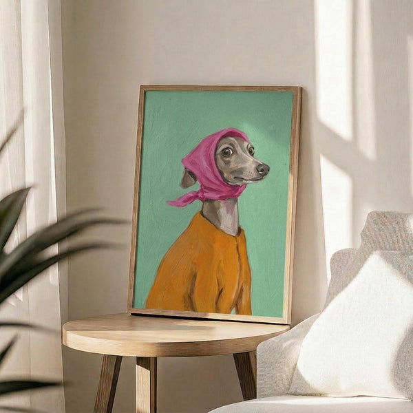 May include: Framed artwork displayed on a round wooden side table next to a beige upholstered chair with white pillow. The painting shows an Italian Greyhound dog wearing a bright pink headscarf and orange sweater against a mint green background. Natural lighting creates soft shadows on white walls, with a plant visible in the corner of the modern, minimalist room setting.