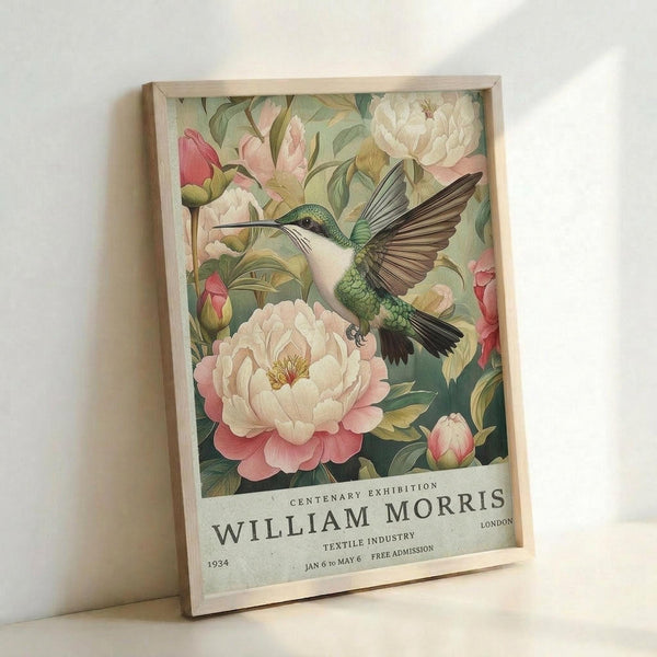 A framed vintage-style poster featuring a detailed hummingbird with iridescent green and blue feathers hovering among large pink and white peonies with lush green foliage. The artwork has a botanical illustration aesthetic with rich, saturated colors against a muted background. Below the floral design, text reads "CENTENARY EXHIBITION WILLIAM MORRIS LONDON TEXTILE INDUSTRY 1934 JAN 6 TO MAY 6 FREE ADMISSION." The poster is displayed in a light wooden frame leaning against a white wall.