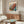May include: Colorful vintage exhibition poster from 1986 featuring abstract botanical design with orange, blue, and teal circular forms resembling flowers or berries. Poster displayed in thin frame above wooden sideboard with sliding doors. Modern white sculptural chair and glass-top oval coffee table complete the mid-century modern living room scene with white walls and natural lighting.