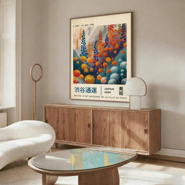 May include: Colorful vintage exhibition poster from 1986 featuring abstract botanical design with orange, blue, and teal circular forms resembling flowers or berries. Poster displayed in thin frame above wooden sideboard with sliding doors. Modern white sculptural chair and glass-top oval coffee table complete the mid-century modern living room scene with white walls and natural lighting.