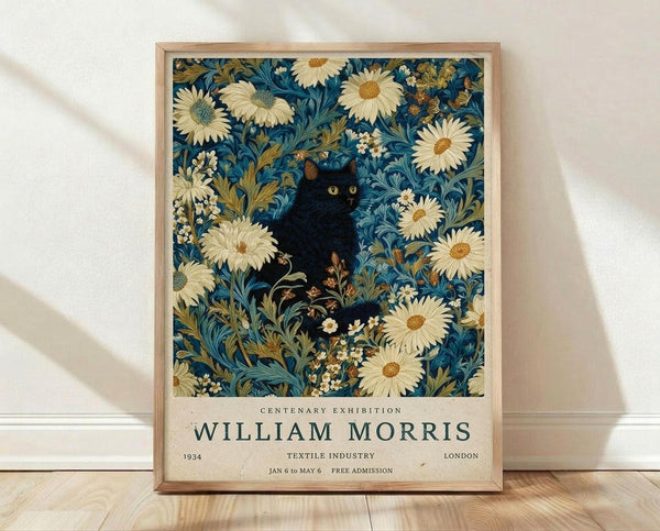 May include: Framed poster print with vintage-style botanical pattern in deep blue background featuring cream-colored daisies with orange centers, ornate foliage, and a black cat with green eyes nestled among the flowers. Exhibition text reads "Centenary Exhibition William Morris 1934 Textile Industry London Jan 6 to May 6 Free Admission." Natural wood frame displayed on light wooden floor against white wall with soft shadows.