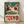 May include: Framed vintage-style poster featuring a stylized frog with large red-rimmed eyes and pointed ears against a teal green background. Bold red "TOKYO" text overlays Japanese characters. The distressed, weathered design has cream and red color accents. Wooden frame sits on light wood flooring against white wall with natural lighting.
