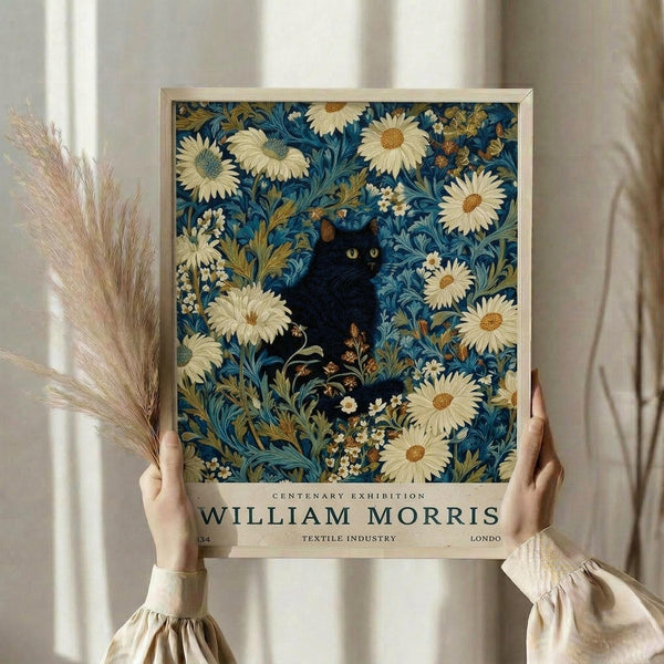 May include: Hands holding a framed art print featuring a black cat with bright yellow eyes surrounded by white daisies and blue-green foliage in William Morris style. The poster has exhibition text at bottom reading "Centenary Exhibition William Morris Textile Industry London." Person wears cream ruffled sleeves and holds dried wheat stems. Neutral curtain background with soft natural lighting.