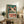 May include: Vintage-style poster featuring a white frog character with large green eyes and red markings, displaying "TOKYO" in bold red letters with Japanese characters above. The teal and cream colored artwork is displayed in a light wood frame, leaning against a white wall on a rustic wooden sideboard alongside potted plants, books, and ceramic vessels in a cozy living room setting with a beige armchair visible.
