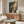 May include: Whimsical poster artwork showing a green frog sitting on a toilet reading a newspaper, surrounded by plants in warm lighting. The illustration is framed in natural wood and displayed above a mid-century modern walnut credenza with sliding doors. Room features a curved white upholstered chair, oval glass coffee table, white table lamp, and brass floor mirror in a minimalist living space with cream walls and natural sunlight.