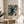 May include: Framed poster featuring a black cat with bright green eyes surrounded by blue floral wallpaper pattern with white daisies and botanical motifs. Exhibition-style print with "William Morris" and "Centenary Exhibition" text at bottom. Wooden frame sits on round wooden side table next to beige upholstered chair. Soft natural lighting creates shadows on neutral wall. Plant leaves visible in corner.