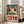 May include: A framed vintage-style poster featuring a whimsical frog character with large round eyes and red spots, displayed in a bright living room setting. The artwork has a distressed teal and cream color scheme with "TOKYO" text and Japanese characters. The wooden-framed print leans against a white wall near large windows, positioned next to a beige sectional sofa on light hardwood floors.