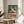 May include: Framed vintage-style poster with teal background featuring a white cartoon frog with large red-rimmed eyes and red spots, displaying "TOKYO" in bold red letters with Japanese characters above. Poster sits on round wooden side table next to beige upholstered chair with white pillow, green plant leaves visible on left, natural lighting creating soft shadows on white wall.