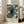 May include: Vintage-style exhibition poster featuring a black cat with yellow eyes surrounded by white daisies and blue-green foliage in William Morris decorative pattern style. Cream text area shows "CENTENARY EXHIBITION WILLIAM MORRIS 1934 TEXTILE INDUSTRY LONDON JAN 6 to MAY 6 FREE ADMISSION." Framed print leans against white wall next to beige upholstered chair in bright, airy room with hardwood floors and large windows.