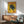 May include: Vintage-style poster featuring a black panther perched on a tree branch against a yellow background, with "TOKYO" in red letters and Japanese characters below, displayed in a wooden frame above a mid-century modern credenza. Room setting shows a mint green upholstered chair with wooden arms, small white table lamp, and round wooden side table, all bathed in natural sunlight creating geometric shadows on white walls.