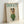 May include: Framed poster print leaning against white wall showing white poodle wearing green knit sweater, patterned yellow scarf wrapped around head, and dark sunglasses against cream background with "DOGUE" text in teal letters at top, displayed in light wood frame