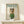 May include: Framed poster showing a white poodle wearing round sunglasses, green knit sweater, and colorful striped headscarf styled like a vintage fashion magazine cover with "DOGUE" in teal letters at top. Light wood frame sits on pale wooden surface next to small white wrapped gift box with natural twine and sprig. Cream colored wall background with soft natural lighting.
