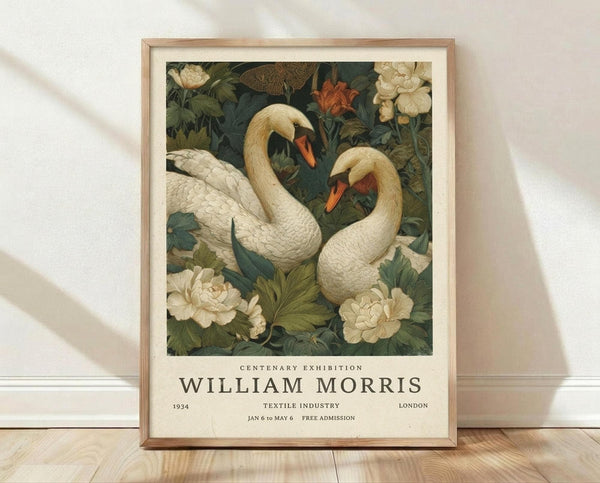 May include: Framed vintage-style poster featuring two white swans with orange beaks surrounded by cream and coral flowers and green foliage. The artwork has a decorative Arts and Crafts movement aesthetic with detailed botanical elements. Below the illustration is text reading "Centenary Exhibition William Morris" with exhibition details. The poster is displayed in a light wood frame, leaning against a white textured wall on light wooden flooring with soft natural lighting creating shadows.