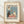 May include: Framed vintage-style poster with cream background featuring two cats sitting at a pink tablecloth-covered table in a blue room. Black cat and white cat with sunglasses share coffee and pastries, with a small flower in vase between them. French text "Café Au Lait" above and "S'il Vous Plaît" below the illustration. Light wood frame rests against white wall on wooden floor with natural lighting casting soft shadows.