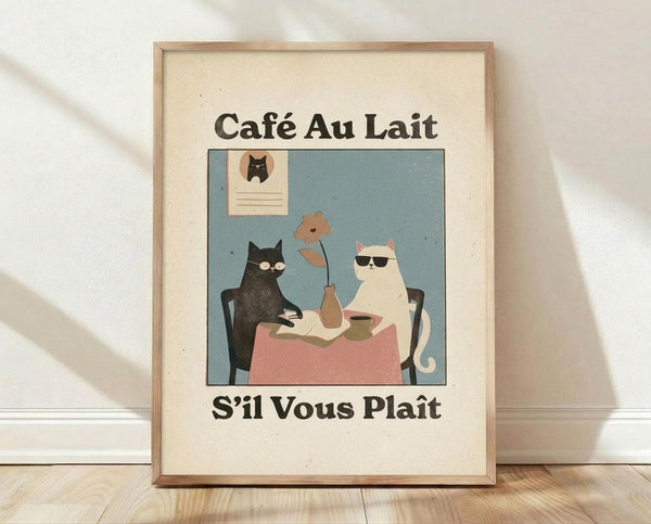 May include: Framed vintage-style poster with cream background featuring two cats sitting at a pink tablecloth-covered table in a blue room. Black cat and white cat with sunglasses share coffee and pastries, with a small flower in vase between them. French text "Café Au Lait" above and "S'il Vous Plaît" below the illustration. Light wood frame rests against white wall on wooden floor with natural lighting casting soft shadows.