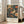 May include: Framed vintage-style poster with colorful bird and floral design in warm tones of orange, cream, and teal, leaning against white wall next to beige upholstered chair in bright room with large windows and natural light streaming across wooden floors.