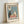 May include: Framed vintage-style poster print leaning against white wall. Features two cats at cafe table - black cat with glasses and white cat with sunglasses - sharing coffee and pastries. Pink tablecloth, blue background, cat portrait on wall. French text "Café Au Lait" and "S'il Vous Plaît" in black letters. Light wood frame, cream colored paper with speckled texture.