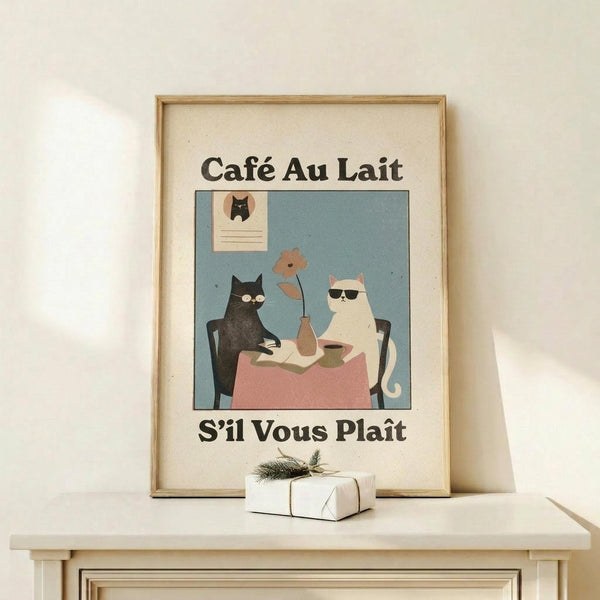 May include: Framed vintage-style poster with cream matting showing two cats at a cafe table - one black cat and one white cat wearing sunglasses, sitting at a pink-clothed table with coffee and a flower vase. French text reads "Café Au Lait" and "S'il Vous Plaît" in black lettering. Light wood frame displayed on white wooden shelf beside small wrapped gift with evergreen sprig against cream wall.