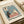 May include: Vintage-style illustrated poster featuring two cats at a cafe table - a black cat and white cat with sunglasses sharing coffee and a flower. Text reads "Cafe Au Lait" and "S'il Vous Plait" in retro typography. Soft blue, pink, and cream color palette with textured paper appearance. Print displayed in white matted frame on neutral surface with ribbon visible.
