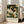 May include: Vintage-style poster featuring two white swans with orange beaks surrounded by cream peonies and lush green foliage on dark teal background. Text reads "Centenary Exhibition William Morris 1934 Textile Industry London Jan 6 to May 6 Free Admission." Framed print leans against white wall next to beige upholstered chair in bright, airy room with large windows.