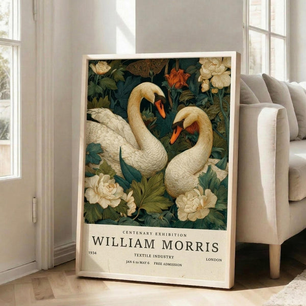May include: Vintage-style poster featuring two white swans with orange beaks surrounded by cream peonies and lush green foliage on dark teal background. Text reads "Centenary Exhibition William Morris 1934 Textile Industry London Jan 6 to May 6 Free Admission." Framed print leans against white wall next to beige upholstered chair in bright, airy room with large windows.