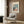 May include: Framed poster print hanging on wood-paneled wall above beige upholstered armchair with geometric pillow. Artwork shows stylized cats at cafe table - black cat and white cat wearing sunglasses sitting with coffee cups on pink tablecloth against teal background. French text "Café Au Lait" and "S'il Vous Plaît" in black letters. Modern reading nook with brass floor lamp and side table.