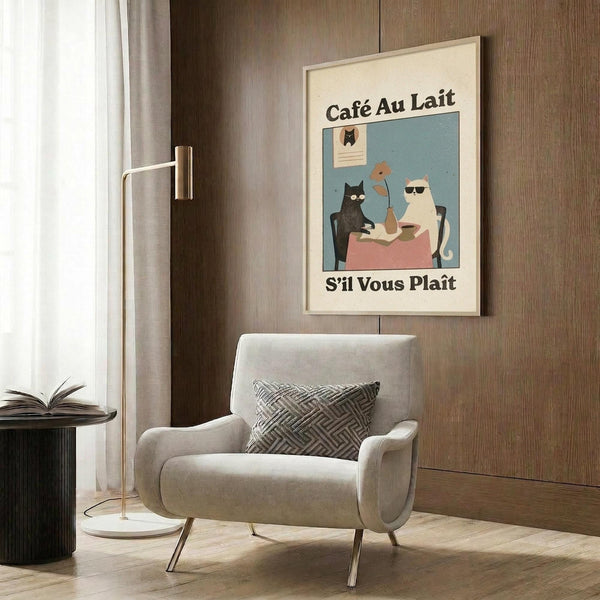May include: Framed poster print hanging on wood-paneled wall above beige upholstered armchair with geometric pillow. Artwork shows stylized cats at cafe table - black cat and white cat wearing sunglasses sitting with coffee cups on pink tablecloth against teal background. French text "Café Au Lait" and "S'il Vous Plaît" in black letters. Modern reading nook with brass floor lamp and side table.