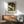 May include: Framed poster featuring two white swans among cream peonies and dark green foliage in William Morris's decorative style, displayed above a mid-century modern wooden credenza with sliding doors. A sage green upholstered armchair with wooden frame sits nearby, along with a small round side table and white ceramic table lamp. Warm natural lighting creates geometric shadows on the white wall.