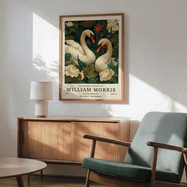 May include: Framed poster featuring two white swans among cream peonies and dark green foliage in William Morris's decorative style, displayed above a mid-century modern wooden credenza with sliding doors. A sage green upholstered armchair with wooden frame sits nearby, along with a small round side table and white ceramic table lamp. Warm natural lighting creates geometric shadows on the white wall.