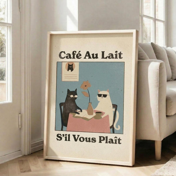 May include: Framed vintage-style poster featuring two cats sitting at a small round table with pink tablecloth, sharing coffee and a flower. Dark cat wears glasses, white cat wears sunglasses. French text reads "Café Au Lait" and "S'il Vous Plaît" against cream background with blue accent. Light wooden frame leans against white wall next to beige upholstered chair in bright, airy room with large windows.