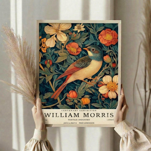 May include: Person holding framed poster featuring colorful bird perched among flowers and foliage in William Morris decorative style. Artwork shows cream and orange blooms with teal-green leaves on dark background. Light wood frame, dried pampas grass visible, neutral clothing and curtained window setting.