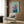 May include: Framed artwork showing an Italian Greyhound wearing a purple head scarf and teal garment against a mint green background, displayed on wood paneled wall above a beige upholstered armchair with geometric patterned pillow. Modern living room setting with brass floor lamp and side table with open book.