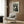 May include: Black and white vintage-style poster featuring three stylized cats with geometric dot patterns, displayed in light wood frame on wood paneled wall above beige upholstered armchair with patterned throw pillow. Modern floor lamp with brass finish and round table with open book visible in contemporary living room setting with neutral tones.