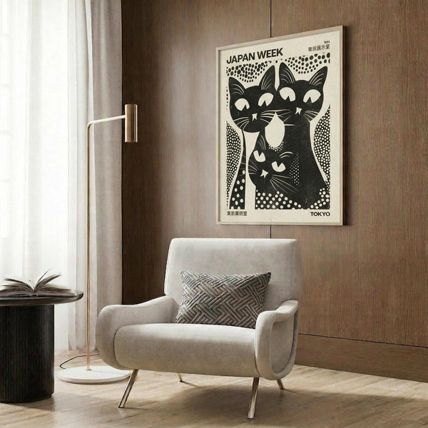 May include: Black and white vintage-style poster featuring three stylized cats with geometric dot patterns, displayed in light wood frame on wood paneled wall above beige upholstered armchair with patterned throw pillow. Modern floor lamp with brass finish and round table with open book visible in contemporary living room setting with neutral tones.