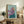 May include: Framed painting of an Italian Greyhound wearing a flowing purple headscarf and teal garment against a soft blue-green background, displayed on a rustic wooden console table alongside ferns, books, and ceramic pottery in a cozy living room setting with neutral tones and natural lighting.