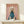 May include: Framed poster showing a white poodle wearing a teal blue turtleneck sweater against a soft pink background, with "DOGUE" text in orange letters at top. Gold frame sits on light wood shelf next to small wrapped white gift box with evergreen sprig. Clean white wall background with natural lighting creates shadow on wall.